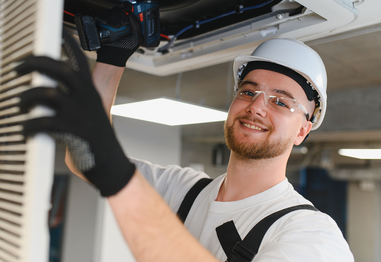 Monteur mit Helm und Schutzbrille bei Klima-Reparatur — Diagnose am Decken-Innengerät